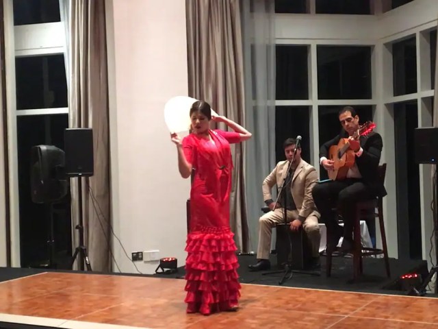 Flamenco dancer in a red ruffled dress performing with live musicians on an indoor wooden floor.