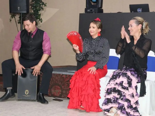 Flamenco trio with a cajón player, a dancer holding a fan and a performer clapping palmas at an indoor event.